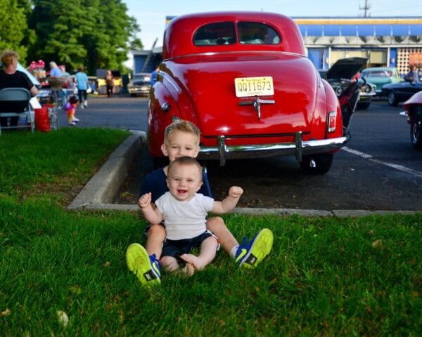 Two young boys sitting in the grass next to a red car.